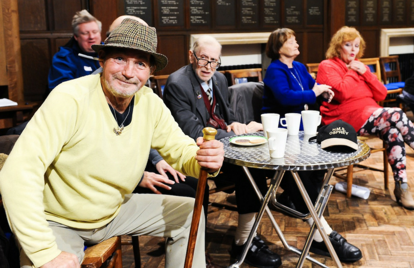 Group of older people having coffee - one man sits holding stick looking directly at camera