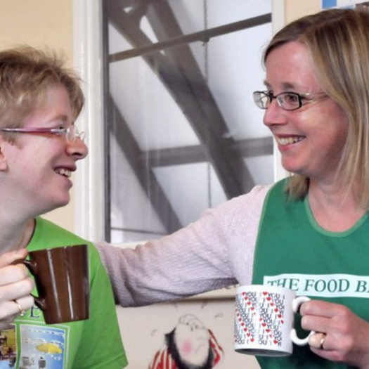 A smiling helper at a food bank is enjoying a hot drink with a visitor to the food bank.
