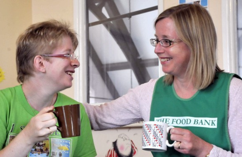 Cost of living news article March23 A smiling helper at a food bank is enjoying a hot drink with a visitor to the food bank.