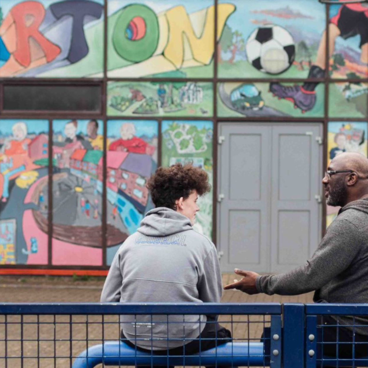 Living Essentials news story 810×525 Teenage boy and man sitting on a bench chatting in front of a mural about the Barton estate in Oxford.