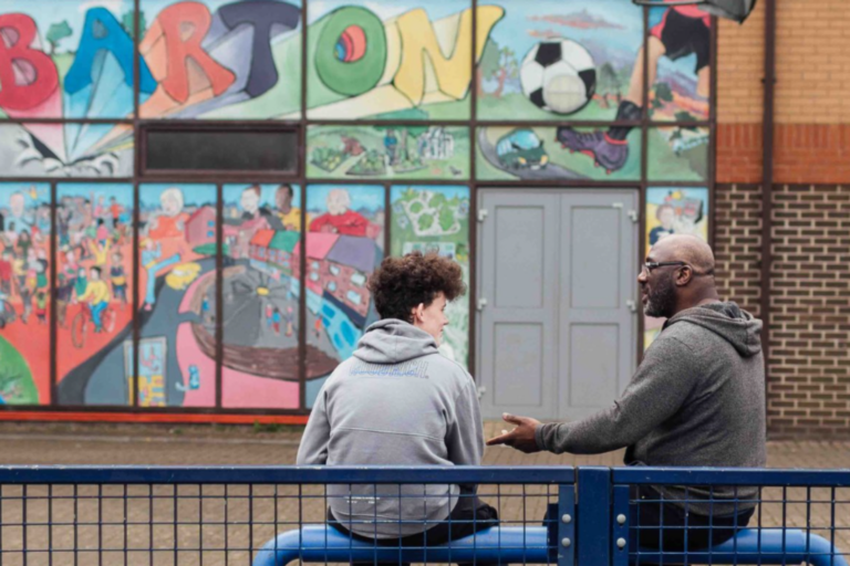 Teenage boy and man sitting on a bench chatting in front of a mural about the Barton estate in Oxford.