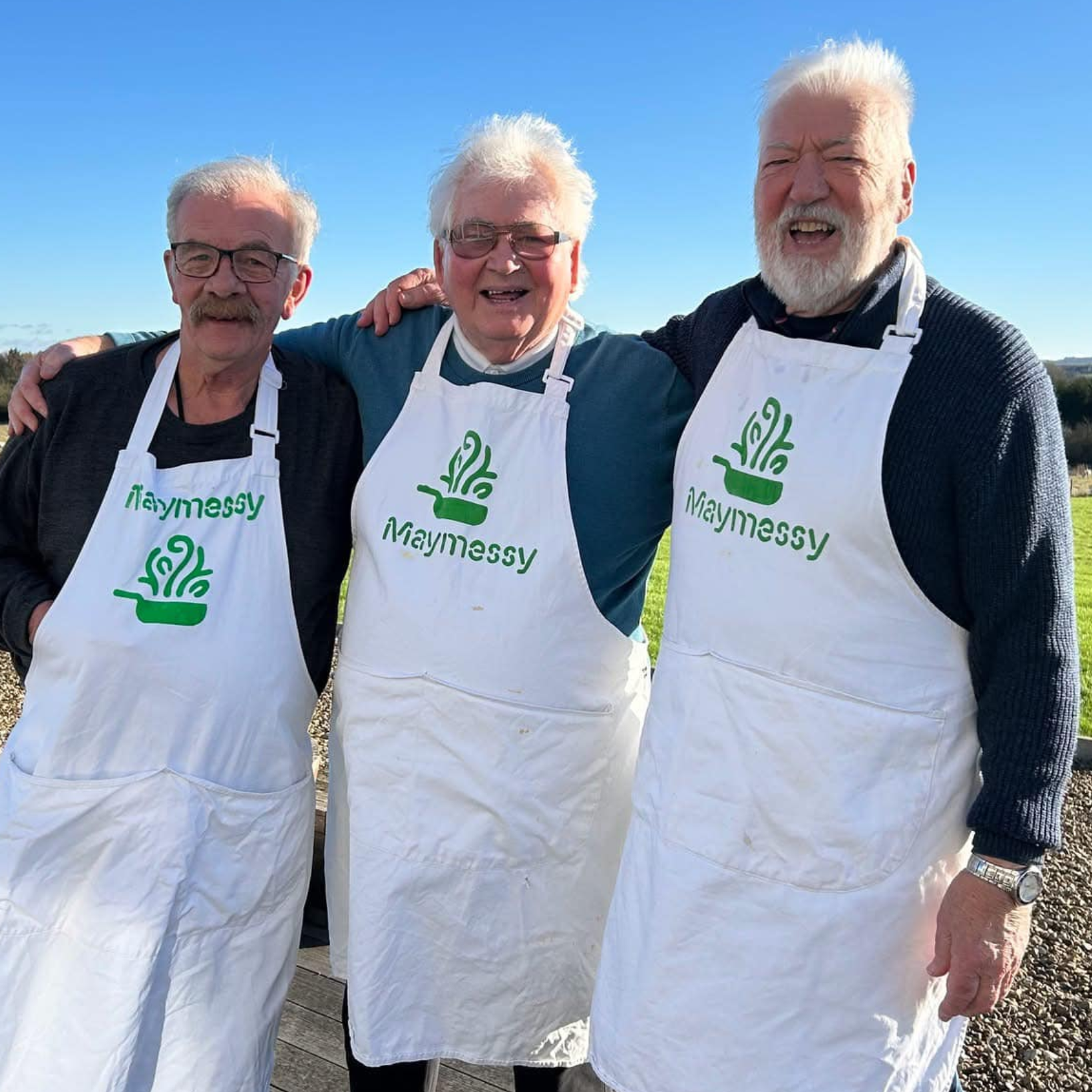 Maymessy Three gentlemen wearing cooking aprons standing with their arms around each other and smiling.