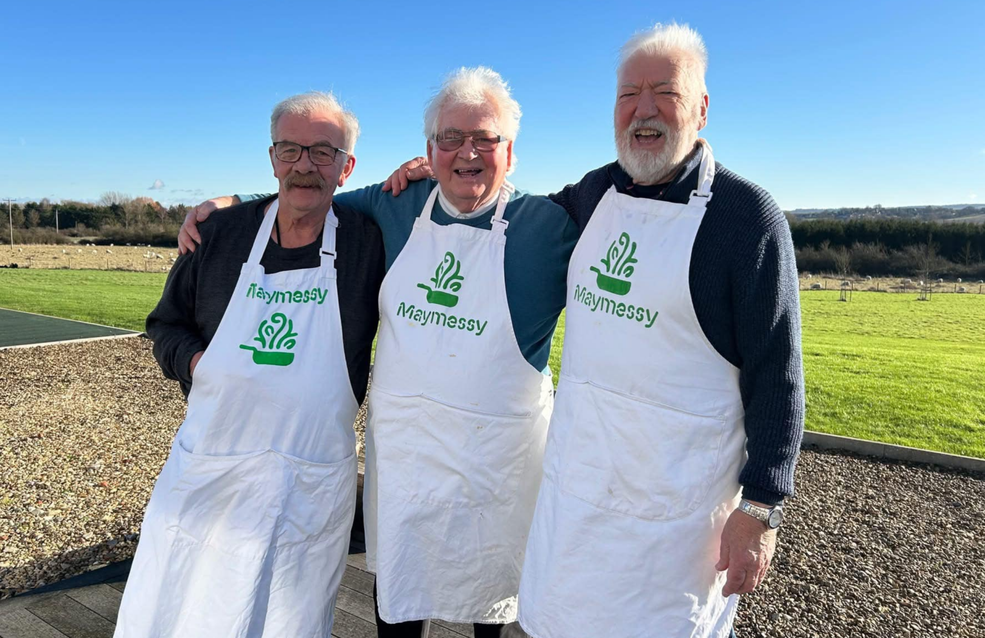 Maymessy Three gentlemen wearing cooking aprons standing with their arms around each other and smiling.
