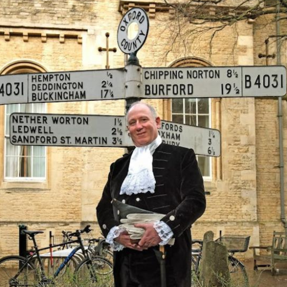 John May news story 810×525 John May, High Sheriff,, standing outside a traditional road sign wearing a black velvet jacket, ruffle shirt, ceremonial sword and white gloves.