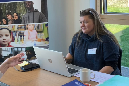A woman sitting in front of a laptop chatting to another woman across the desk.