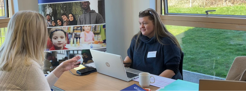 Untitled design (10) A woman sitting in front of a laptop chatting to another woman across the desk.