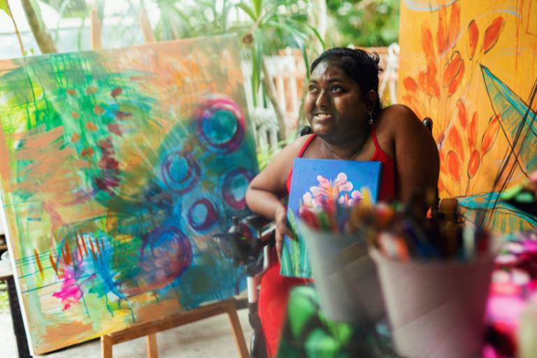 OYAP news story 810×525 Young female person sitting in a wheelchair and painting a picture in an art studio.