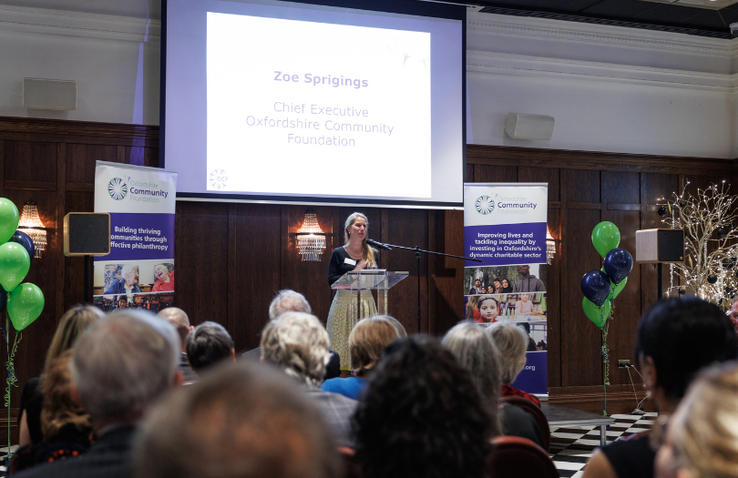 Person standing on stage giving speech with banners and balloons on either side.