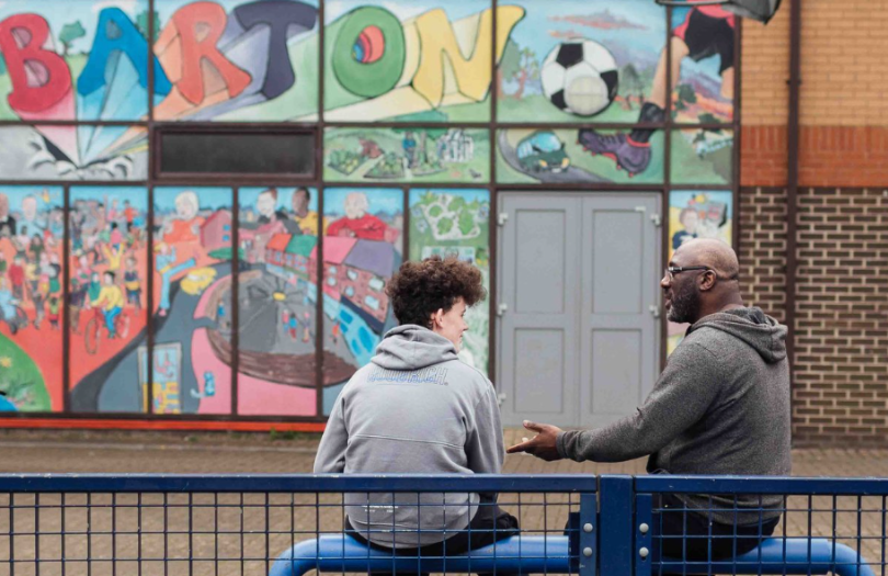 An older black man talks supportively to a younger white man in front a of mural in Barton, Oxford
