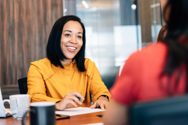 Two women sitting either side of a desk having a conversation while drinking coffee.