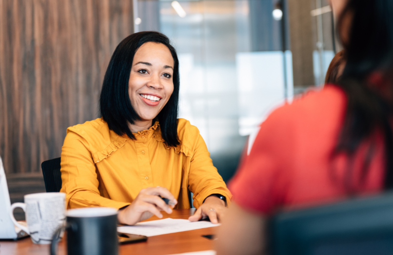 Two women sitting either side of a desk having a conversation while drinking coffee.