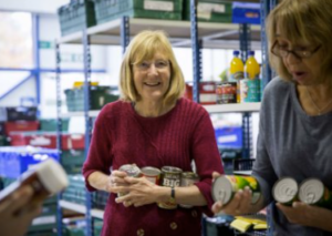 A lady in front of shelves storing food, she is holding some tins of food and smiling.