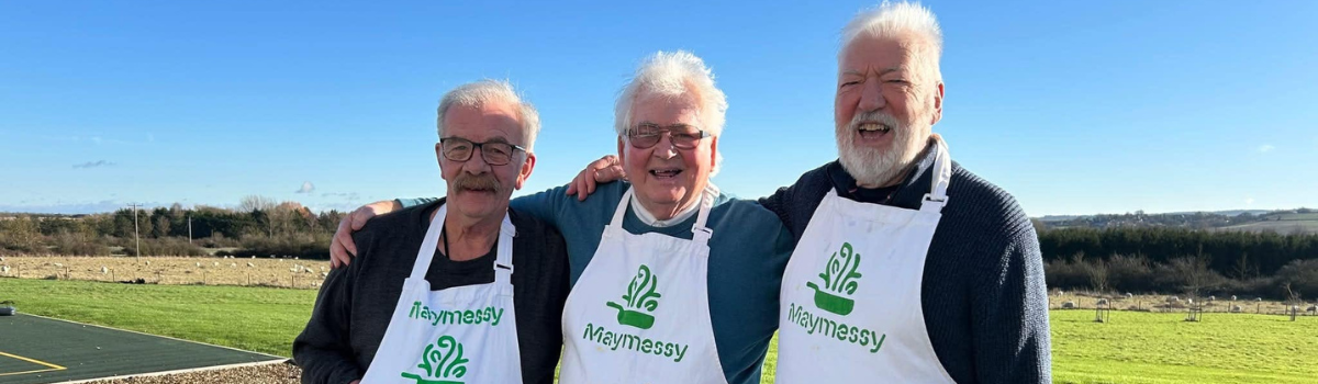 Three men standing outside, wearing cooking aprons and with their arms around each other.