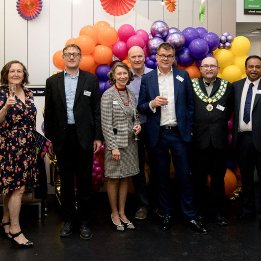 A group of people smiling and holding glasses of fizz in front of balloons and a Didcot Powerhouse banner.