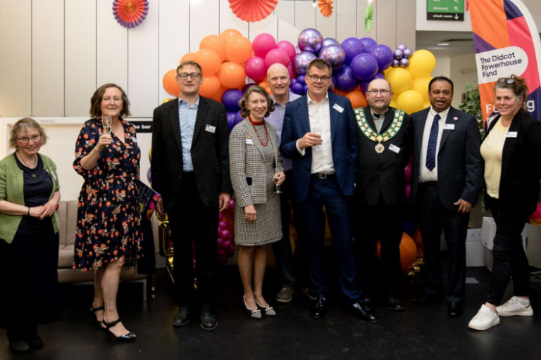 A group of people smiling and holding glasses of fizz in front of balloons and a Didcot Powerhouse banner.
