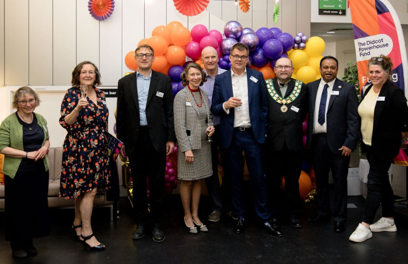 A group of people smiling and holding glasses of fizz in front of balloons and a Didcot Powerhouse banner.