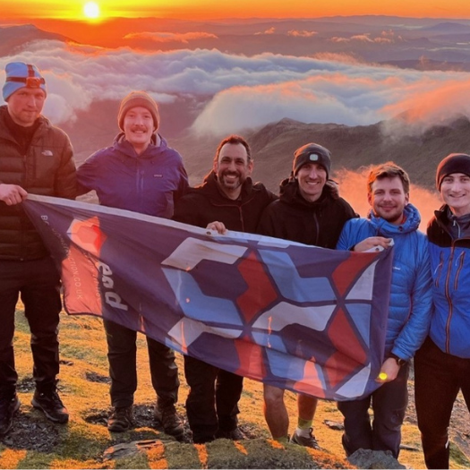 Six people at the top of Snowdon holding a Beard Construction flag with the sun rising behind them.