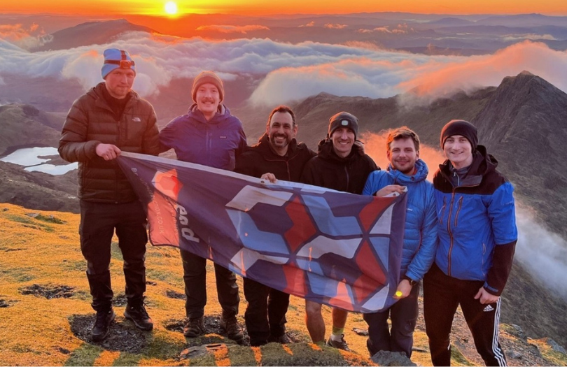 Beard news story Jul 26 810×525 Six people at the top of Snowdon holding a Beard Construction flag with the sun rising behind them.